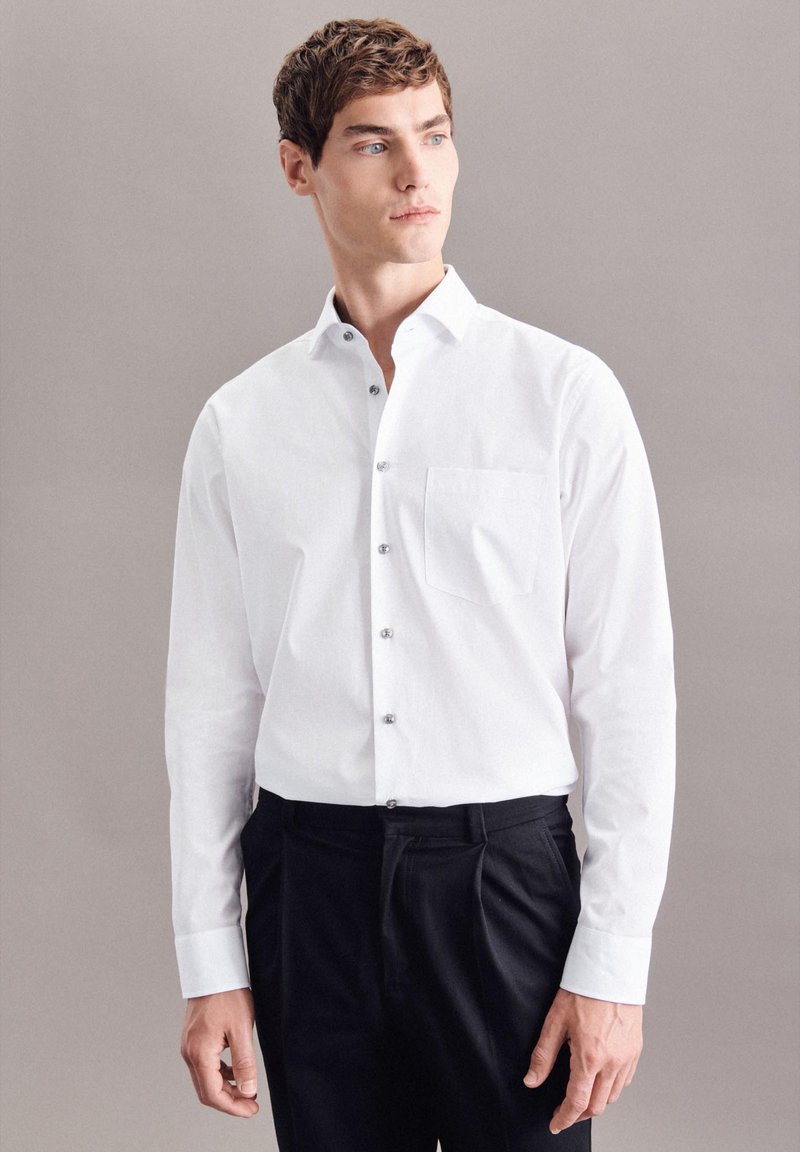 Witte blouse met lange mouwen, een button-down kraag, een borstzak en zilveren knopen. Draag het met zwarte broek tegen een neutrale achtergrond.