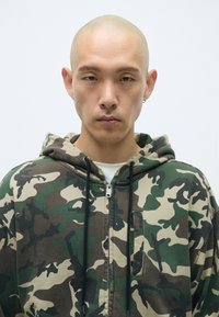 Kameleonu raksta zip-up kapucis ar zaļām, brūnām un bēšām krāsām; iekļauj regulējamu kapuci un mazu priekšējo kabatu.