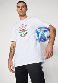 Wit katoenen T-shirt met Arsenal-logo, korte mouwen, losse pasvorm. Zwarte sportbroek met zijstrepen. Blauwe en rode voetbal onder de arm gehouden.