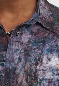 Camicia con bottoni, dotata di un motivo floreale nei toni del viola, blu e verde. Presenta un colletto e un dettaglio decorativo in metallo argentato.