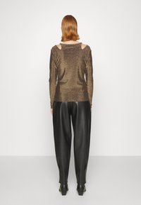 Bronzefarbenes Longsleeve-Shirt mit Schulterauschnitten, kombiniert mit schwarzen, weiten Hosen aus Kunstleder und ankle boots mit Blockabsatz. Glatte Textur.
