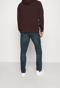 Sudadera burdeos con bolsillo canguro, combinada con jeans de denim azul que cuentan con cinco bolsillos. Modelo lleva zapatillas blancas con acento de logo.