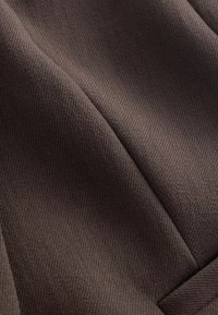 Primo piano di tessuto marrone scuro piegato con trama finemente strutturata e cucitura visibile che attraversa diagonalmente la superficie.