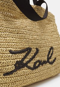 Sac fourre-tout en paille tissée beige avec des poignées en tissu noir et un texte brodé noir sur le devant indiquant "Zola".