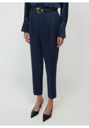 Mujer que lleva pantalones ajustados azul marino, tacones slingback negros con punta afilada, un cinturón negro con hebilla dorada y una blusa azul marino de manga larga.