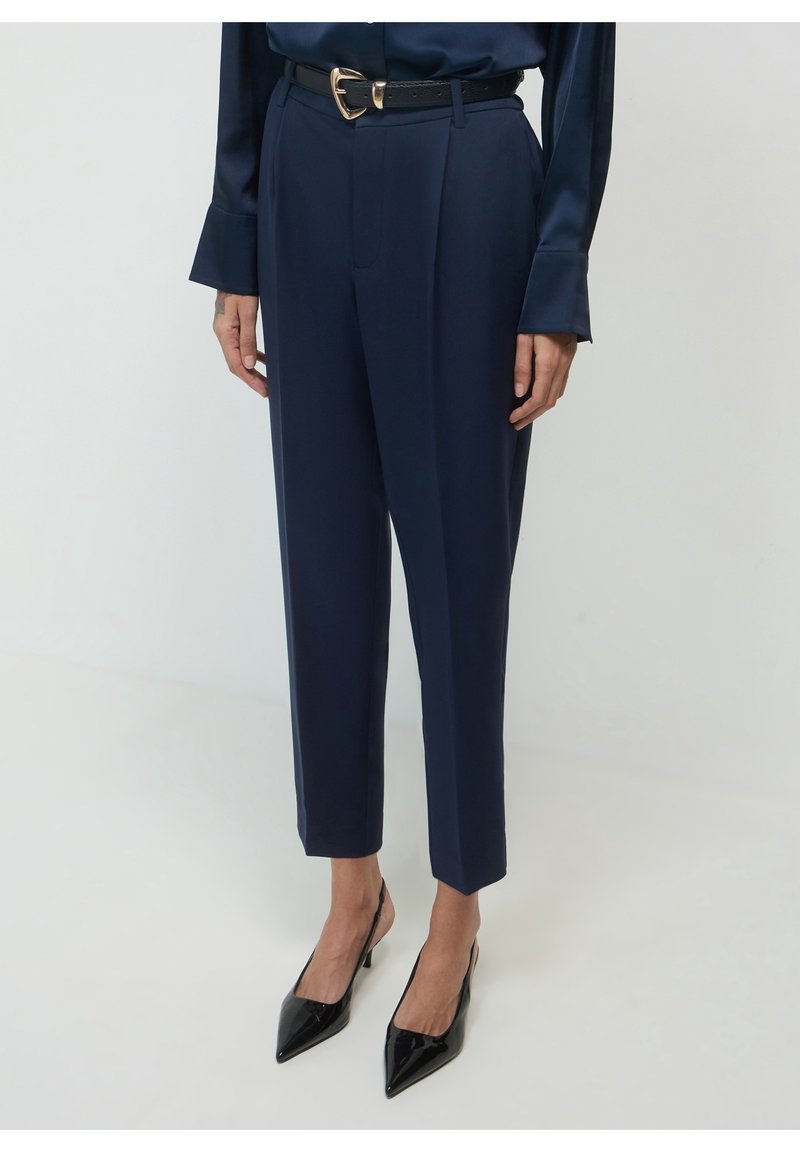Femme portant un pantalon bleu marine sur mesure, des talons slingback noirs à bout pointu, une ceinture noire avec une boucle dorée et un chemisier bleu marine à manches longues.