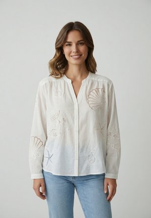 Femme souriante aux cheveux mi-longs portant une chemise blanche à boutons avec des motifs découpés sur le thème de la mer et un jean bleu clair.
