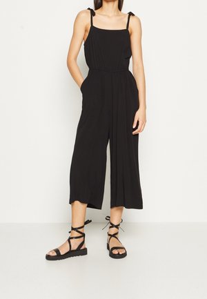 Kvinde iført sort ærmeløs jumpsuit med brede ben og bindebånd samt sorte snøresandaler med flad sål, stående mod en ensfarvet lys væg.