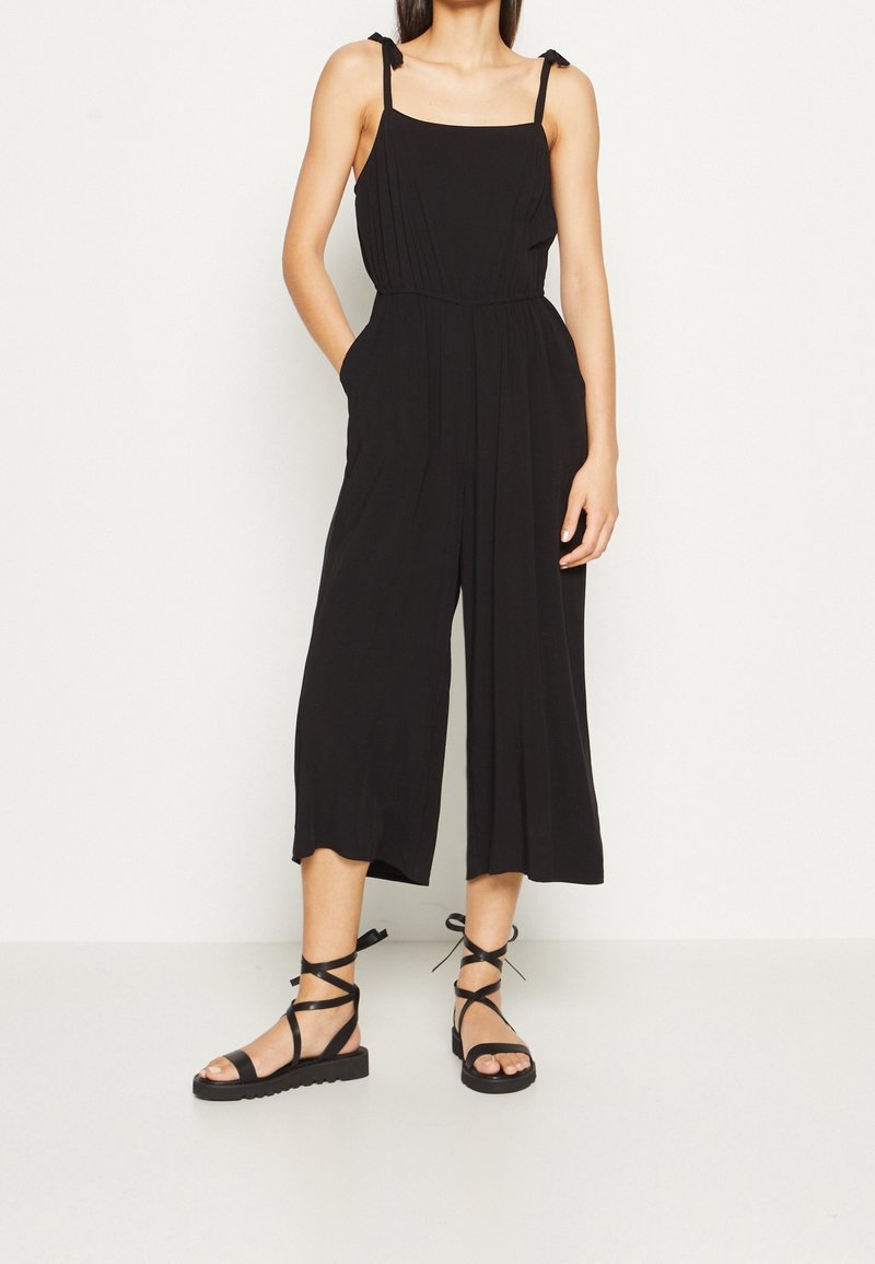 Femme portant une combinaison noire sans manches à jambes larges avec des bretelles à nouer et des sandales plates noires à lacets, debout contre un mur clair uni.