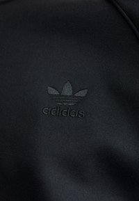 Juodas sportinis audinys su iškilnia Adidas logo, kuriame pavaizduota trijų lapų dizainas, pagerina paviršiaus tekstūrą ir prekės ženklo matomumą.