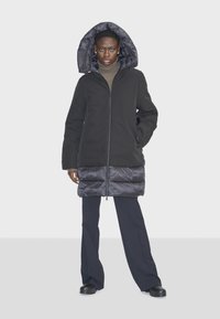 Čierny prešívaný kabát s lesklou spodnou časťou a hrubou podšívkou kapucňou, kombinovaný s tmavými širokými nohavicami a členkovými čižmami.