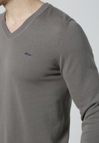 Grauer V-Ausschnitt-Pullover aus gestricktem Material. Verfügt über ein dezentes Logo auf der Brust und gerippte Kanten. Die taillierte Passform betont einen lässigen Stil.
