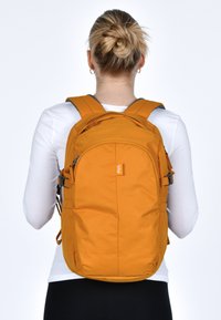 Zaino in tessuto arancione con spallacci imbottiti, più scomparti e una piccola etichetta con logo. Texture liscia e forma arrotondata, adatto per l'uso quotidiano.