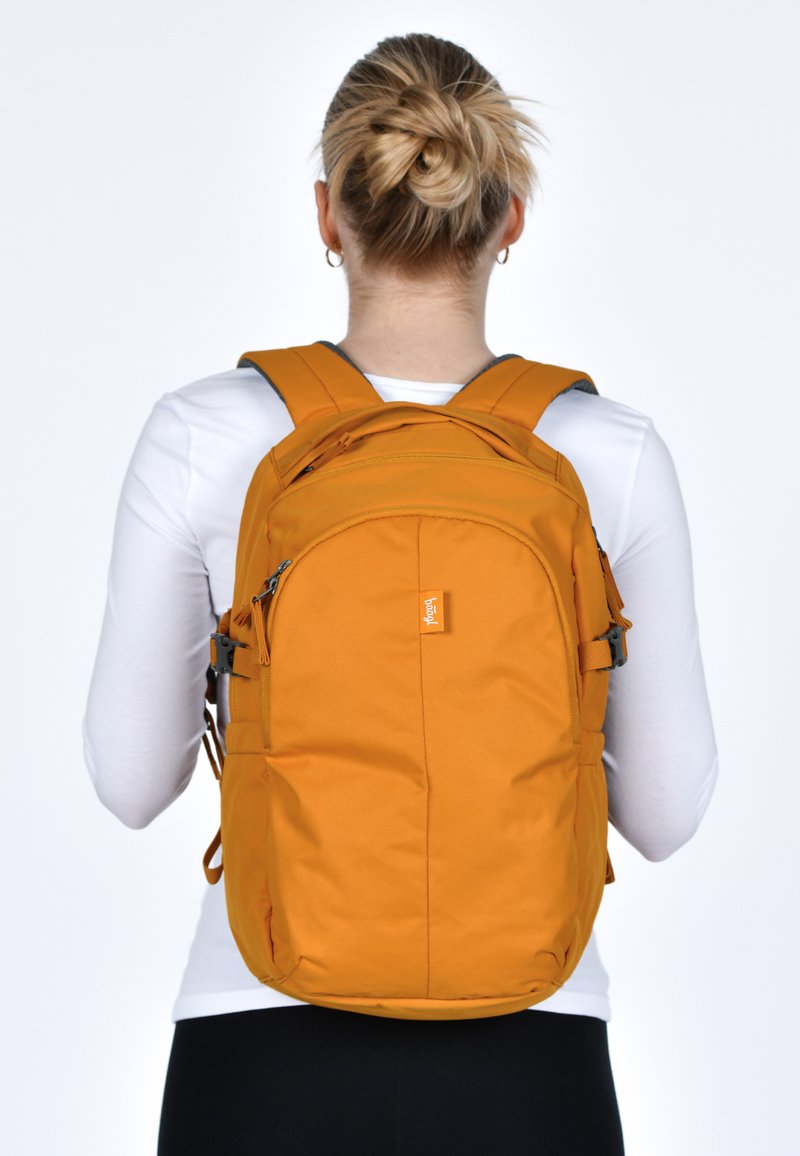Zaino in tessuto arancione con spallacci imbottiti, più scomparti e una piccola etichetta con logo. Texture liscia e forma arrotondata, adatto per l'uso quotidiano.