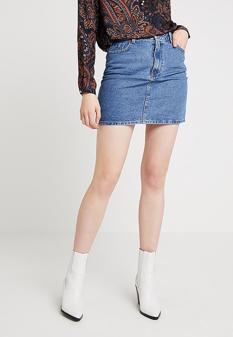 Minifalda de mezclilla con un diseño ajustado, color azul claro y bolsillos frontales. Combinada con botas de tobillo blancas y una blusa negra con estampado.