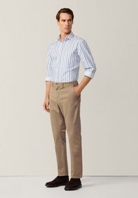 Camicia a righe azzurro chiaro e bianco, abbinata a pantaloni chino beige. Il modello sta in piedi con una mano in tasca, indossando scarpe marroni scure.