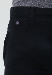 Svarta bomullsbukser har en figurnära design, en synlig knapp med röda och blå accenter, samt en mjuk textur. Midjan är strömlinjeformad.