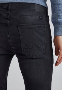 Svarta denimjeans har en bakficka med en subtil broderad bergsdesign och en strukturerad midjebandsetikett. Materialet verkar mjukt och hållbart.