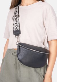 Schwarze Leder-Umhängetasche mit strukturierter Oberfläche, mit Reißverschluss und gemustertem, verstellbarem Tragegurt mit geometrischen Motiven.