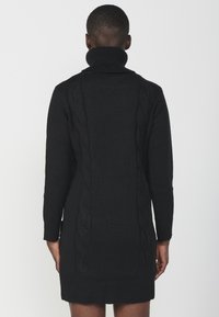 Abito nero lavorato a maglia con alto colletto a cuore, maniche lunghe e dettagli a trecce lungo i lati. Texture liscia e forma aderente.