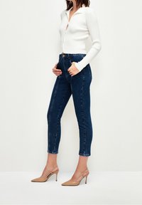 Top blanco con textura de canalé y botones, combinado con jeans de denim oscuro de tiro alto y tacones nude de punta. Los jeans tienen bolsillos frontales y costuras.