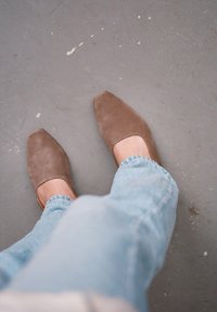 Bruna mockakängor med fyrkantig tådesign, parat med uppvikt ljusblå jeans på en grå yta.