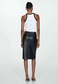 Femme debout de dos, portant un haut blanc sans manches avec un bord noir, une jupe midi en cuir noir et des talons noirs.