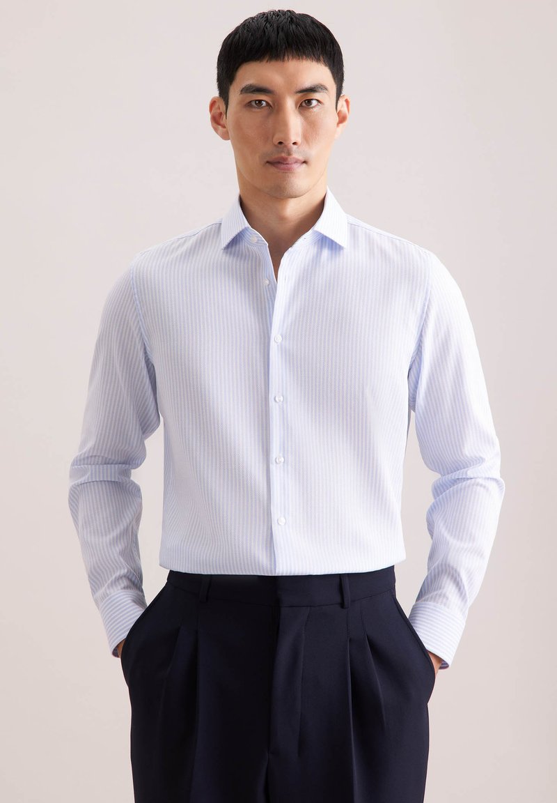 Chemise rayée bleu clair avec col boutonné, manches longues et boutons blancs, associée à un pantalon sombre. Texture de tissu lisse.