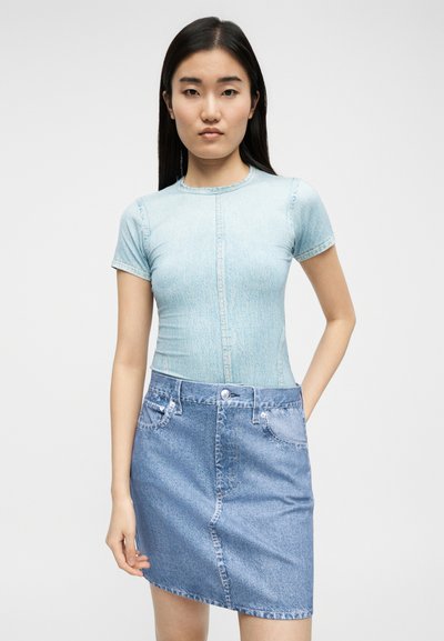 Νεαρή γυναίκα με ίσια μαύρα μαλλιά που φοράει ένα κοντομάνικο ανοιχτό τζιν τοπ και μια ψηλόμεση μπλε τζιν φούστα, στέκεται μπροστά σε λευκό φόντο.