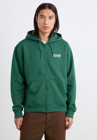 Groene rits hoodie met een voorzak, gemaakt van zachte stof; heeft een logo op de linkerkant van de borst en geribbelde manchetten en onderrand.