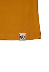 Hem eines senfgelben Baumwoll-T-Shirts mit einem genähten Etikett, das gekreuzte Scheren und die Buchstaben "B" und "R" auf einem weißen Hintergrund zeigt.