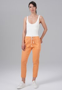 Witte bodysuit met een ronde hals, gecombineerd met oranje joggerpants met trekkoord, voorzien van twee zijzakken en een aansluitende pijpdesign. Witte sneakers.