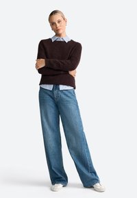 Pull en tricot marron foncé sur une chemise rayée bleu clair, associé à un jean bleu à large jambe et des chaussures décontractées blanches.