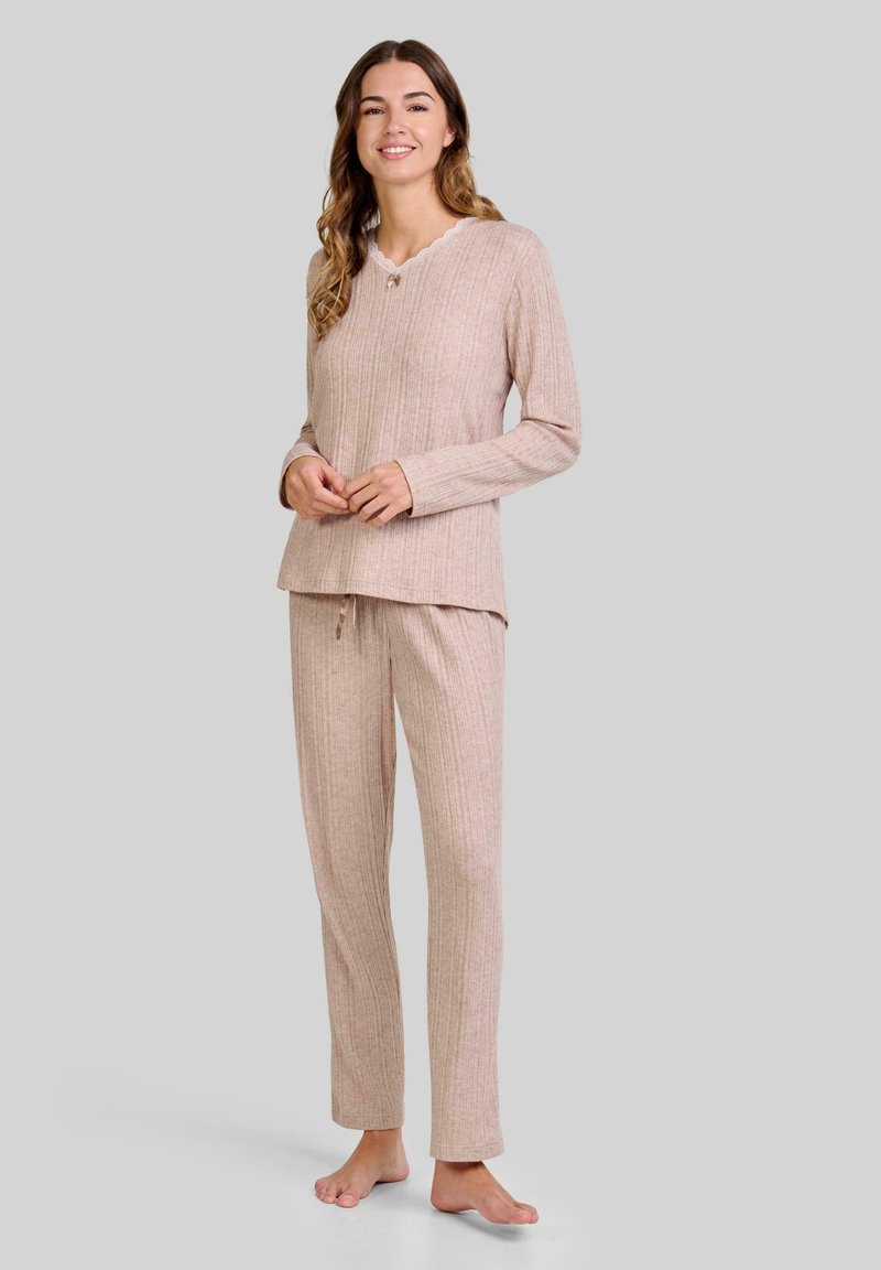 Mujer de pie descalza, vistiendo una blusa de manga larga beige de canalé y pantalones a juego, sobre un fondo gris neutro.