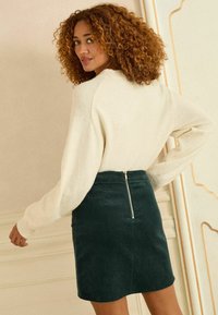 Zelená kordurová sukně se zipem na zádech, zkombinovaná s krémovým svetrem s širokými rukávy. Texturovaný, přiléhavý design.