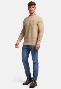 Pull en tricot beige avec des rayures horizontales texturées, associé à un jean bleu et des bottes Chelsea brunes. Le design présente un col rond.