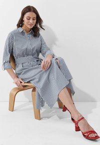 Gestreiftes Hemdkleid in Marineblau und Weiß, mit einem taillierten Gürtel und einem ausgestellten Saum. Kombiniert mit roten Blockabsatzsandalen. Sitzt auf einem Holzstuhl.