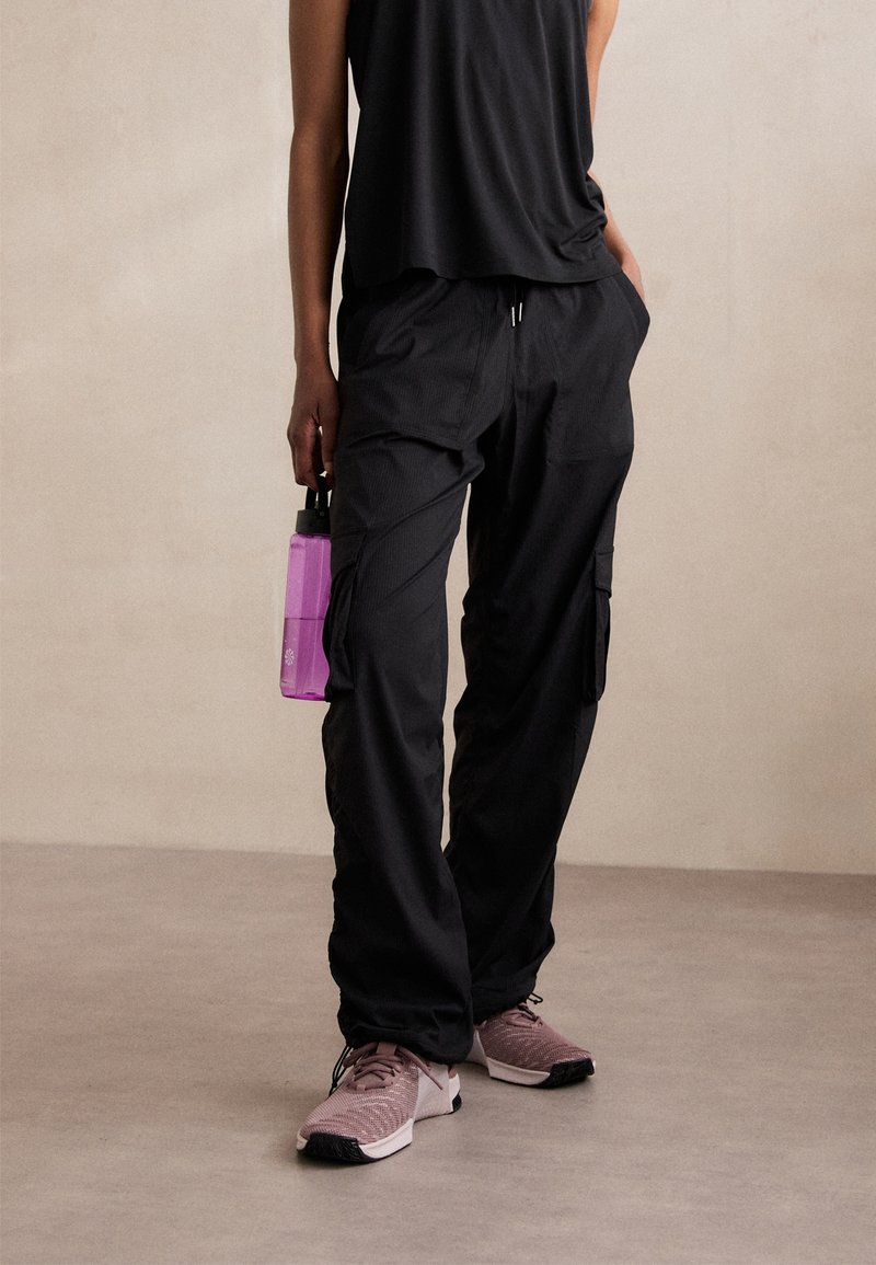 Personne portant un pantalon cargo noir, un haut sans manches noir, des chaussures de course roses, et tenant une bouteille d'eau violette à ses côtés.