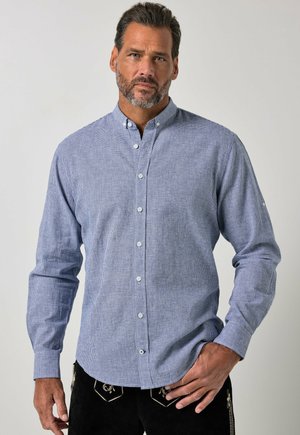 Mand med skæg iført en lyseblå ternet skjorte med knapper og sorte bukser med broderede motiver, stående med den ene hånd i lommen.