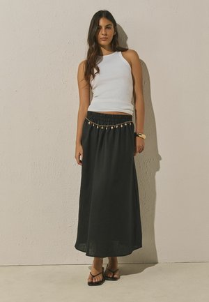 Moteris dėvi baltą be rankovių viršutinę dalį ir ilgą juodą sijoną su auksine grandinės diržu, stovinti prie tekstūruotos smėlio spalvos sienos ir grindų.