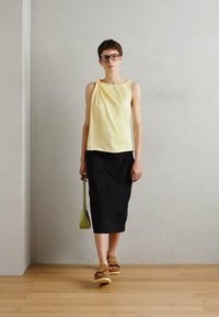 Gulldrapert topp, svart skjørt, brune plattformsandaler og grønn veske. Minimalistisk design med lette materialer og glatte teksturer.
