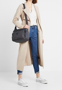 Donna in un trench beige, con una blusa bianca, jeans blu risvoltati, sneakers bianche, che porta una borsa a spalla grigio scuro con tracolla regolabile.