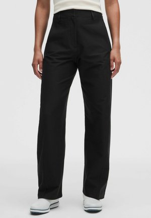 Persona con pantalones negros de tiro alto y corte recto, combinados con zapatos blancos y una camiseta blanca, de pie sobre un fondo liso.