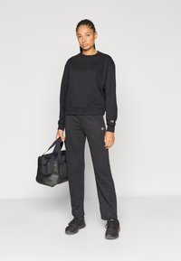 Μαύρο φούτερ και παντελόνι φόρμας από βαμβακερό μείγμα, με ριμπ μανσέτες και λογότυπο. Το μοντέλο κρατάει μια μαύρη τσάντα duffle.