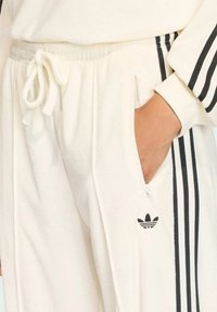 Bílé fleecové tepláky s černými bočními pruhy, elastickým pasem a bočními kapsami. Obsahují vyšívané logo na bocích.