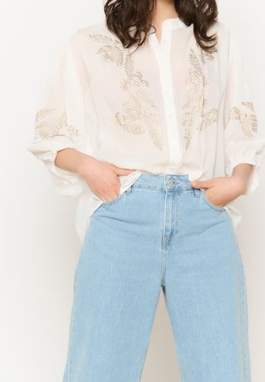 Blouse blanche avec broderie florale dorée, devant boutonné et manches amples. Associée à un jean à jambes larges bleu clair taille haute.