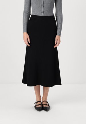 Kobieta ubrana w szary prążkowany top z długim rękawem, czarną spódnicę midi i czarne szpiczaste buty z podwójnymi paskami, stojąca na białej podłodze.