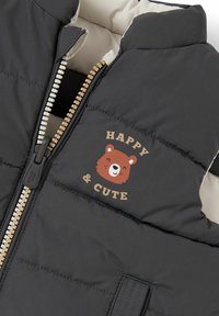 Czarna puchowa kamizelka z beżowym wykończeniem, wyposażona w złoty zamek błyskawiczny i grafikę niedźwiedzia z napisem "SZCZĘŚLIWY I SŁODKI". Miękka, pikowana tekstura.