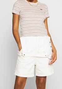 Pruhované, krátké rukávy, růžové tričko s červeným logem, spárované s bílými džíny šortkami. Šortky mají patentky a boční kapsy.