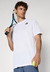 Weißes sportives T-Shirt mit einem Rautenmuster, das ein schwarzes Logo auf der Brust trägt. Das Modell hält einen Tennisschläger und einen Ball.